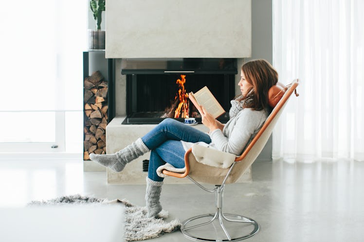 A young woman reads a book while sitting in a chair next to a fire in a winter outfit.