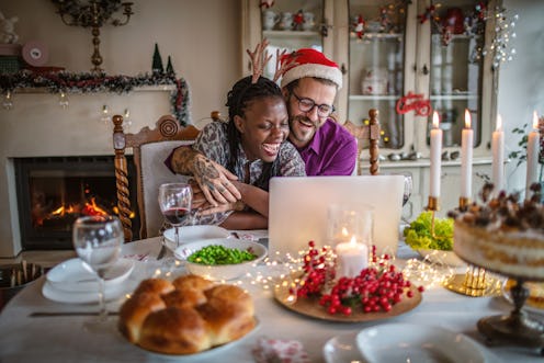 A couple laughs while celebrating the holidays with their laptop on the dinner table. COVID hasn't c...