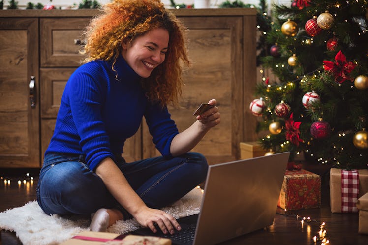 A happy woman shops online next to her Christmas tree and presents.