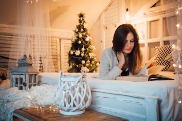 A woman reads a book on her bed in a room decorated for Christmas. 