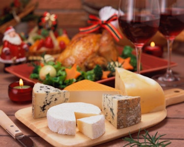 A holiday cheese board is placed on a table next to red wine and a turkey.