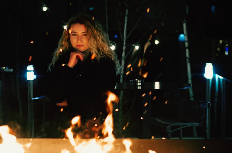 A young woman with blonde hair sits behind a fire pit as embers rise to the night sky.