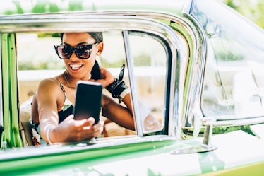 A fashionable woman in sunglasses snaps a selfie in a green vintage car on a sunny day.