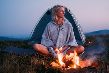 A young woman with blonde hair sits next to a campfire and tent on a grassy mountain.