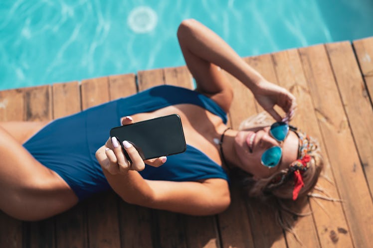 A blonde woman in blue sunglasses, a blue bathing suit, and red hair scarf lays down next to a pool ...