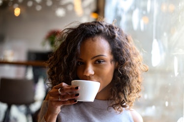 A young Black woman with short, curly hair sips on a cup of espresso while sitting in a cafe.
