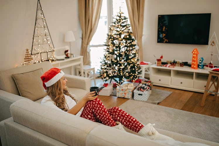 A woman dressed up in Christmas pajamas, watches a movie on the TV in her decorated living room with...