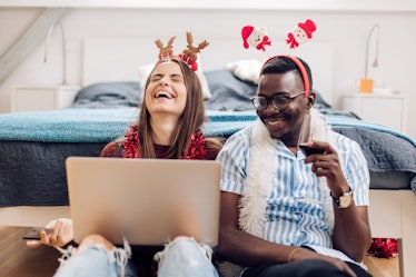 Two happy friends wear festive accessories while laughing and looking at a laptop on Christmas.