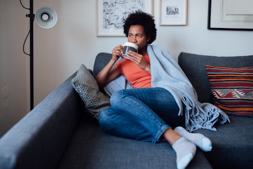 A woman drinks tea on a couch while wearing a blanket. Here are ways to cope with post-election stre...