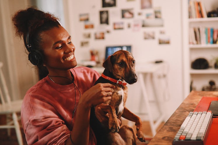 A young Black woman sits at her desk in her home office in a rose-colored crewneck, while holding he...