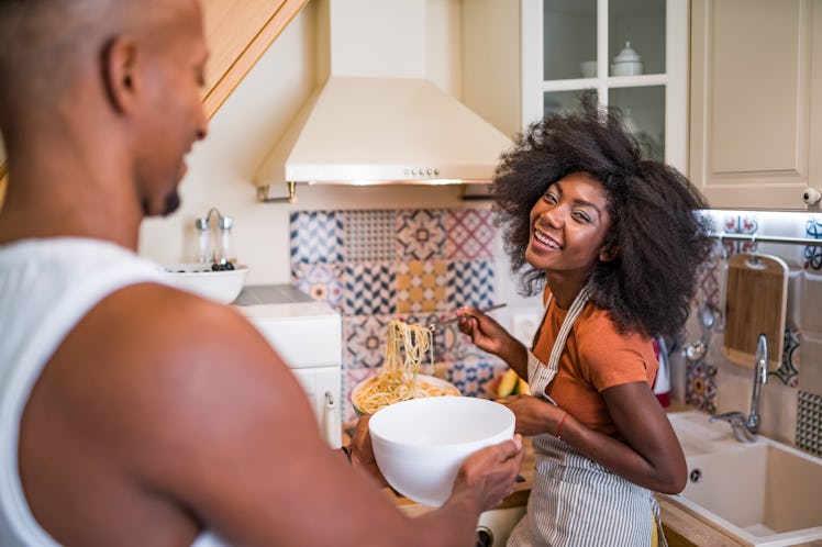 A young Black woman laughs while spooning pasta into a bowl that her partner is holding.