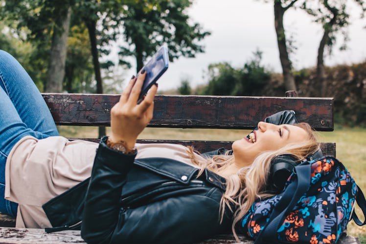 A blonde woman laughs while laying down on a bench on a sunny day in a park.