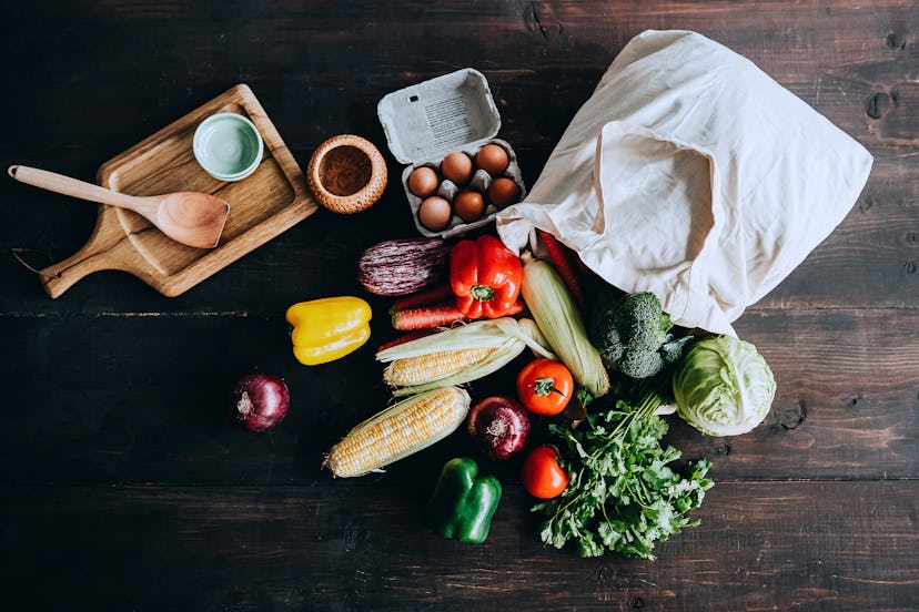 A variety of veggies, a wooden spool, a white bowl and a charcuterie board on a dark wooden surface