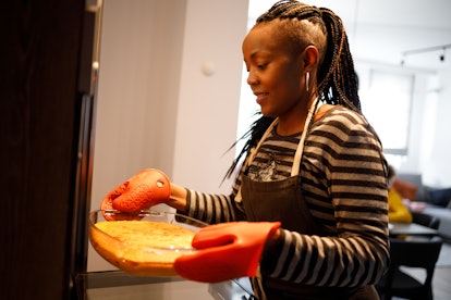 Young lady with dreadlocks putting a glass pan in the oven for her Zoom Thanksgiving meal