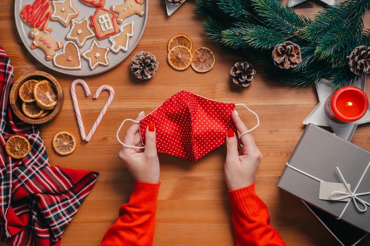 A woman holds a Christmas face mask, while sitting at a table with gingerbread cookies, a present, a...