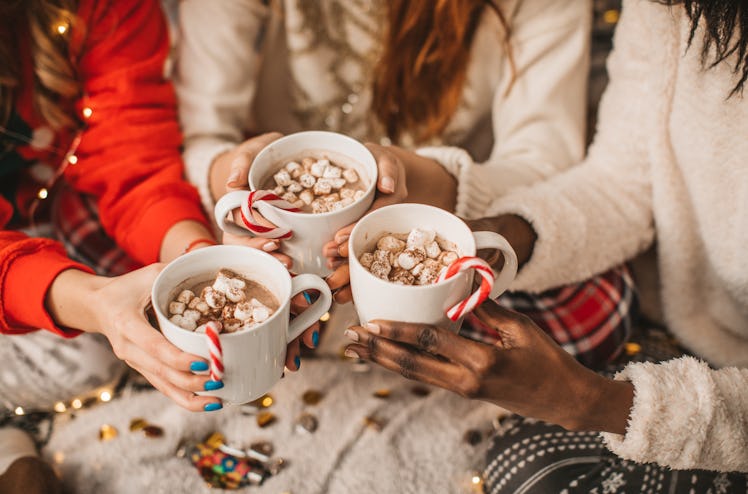 A group of friends holds mugs of hot cocoa, candy canes, and marshmallows together while sitting on ...