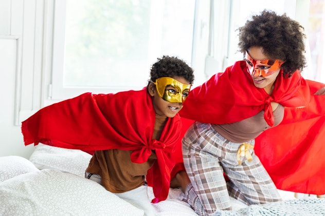 A young boy and a woman in pajamas, a superhero-style eye mask, and red capes, playing on a bed.