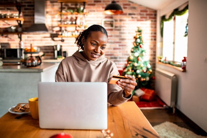 A young Black woman sits at her kitchen table and shops a virtual holiday market on her laptop.