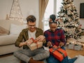 Two housemates sit on their living room floor near a Christmas tree and exchange gifts.