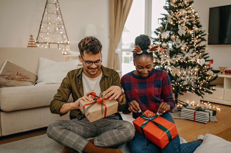 Two housemates sit on their living room floor near a Christmas tree and exchange gifts.