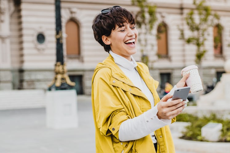 A happy woman holding a cup of coffee in one hand and her phone in the other laughs at something on ...