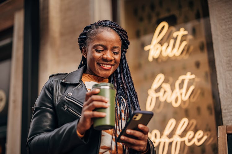 A happy woman in a leather jacket holds a to-go cup of coffee while texting.
