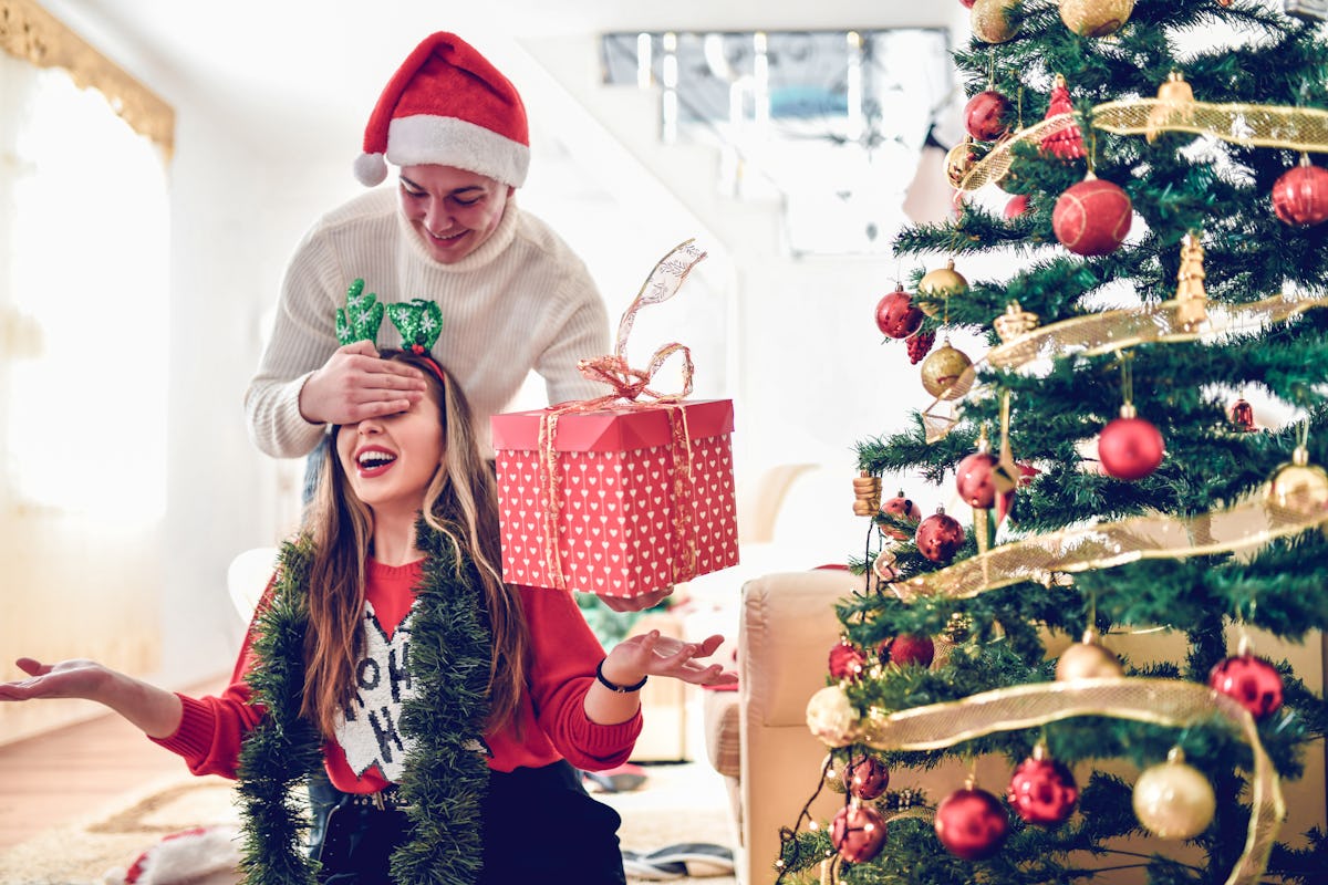 A guy surprises his female friend with a Christmas present, surrounded by Christmas decor in a bright living room.