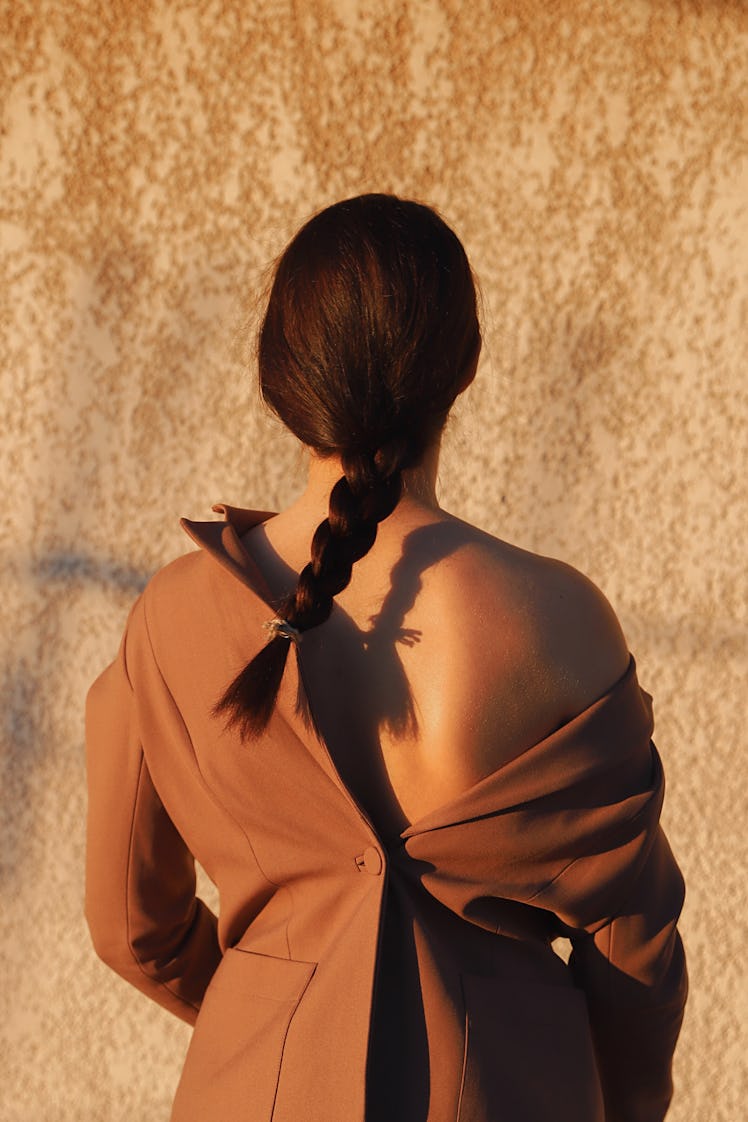 A young woman stands outside in a brown jacket with her hair in a single braid.