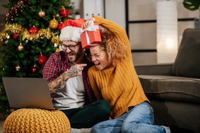 A happy couple poses in front of their laptop while doing a virtual white elephant gift exchange for...