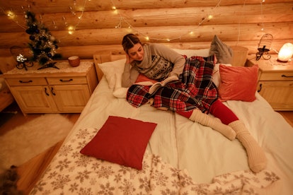 A girl in her Christmas pajamas lounges in bed while reading, laying next to her little Christmas tr...