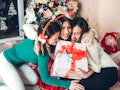 Three friends embrace, holding a Christmas present in front of a Christmas tree and lots of confetti...