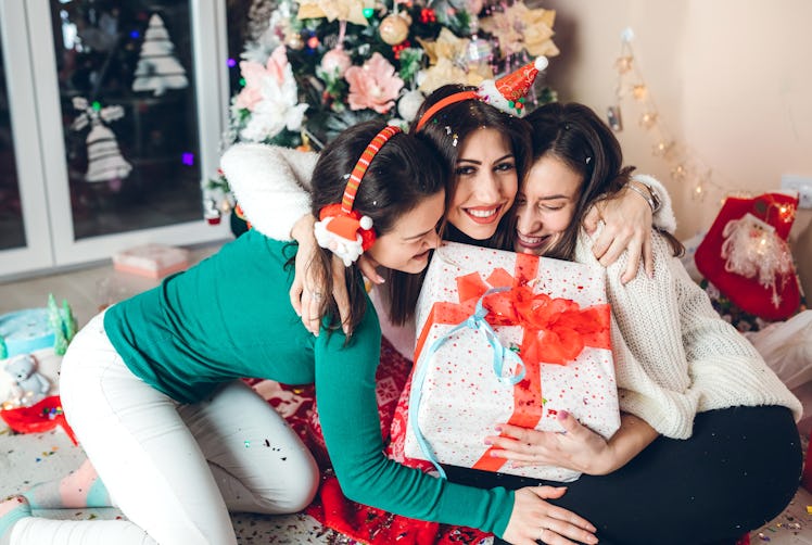Three friends embrace, holding a Christmas present in front of a Christmas tree and lots of confetti...