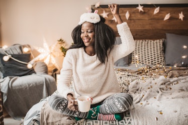 A happy woman dressed in holiday loungewear enjoys a mug of hot cocoa on her bed.
