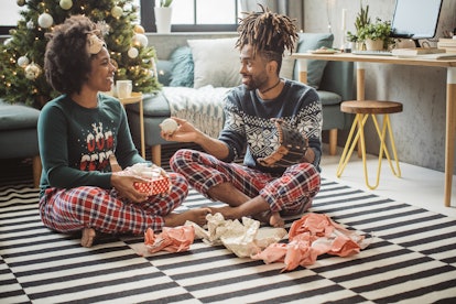 A happy couple wearing Christmas loungewear sits on the floor of their living room while opening pre...