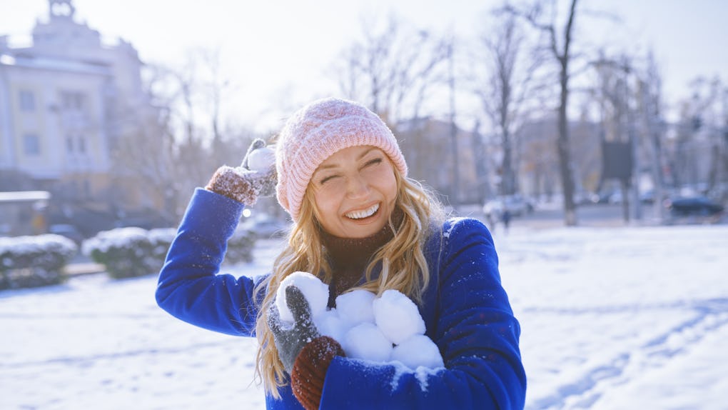 A happy blonde woman in a blue pea coat holds snowballs on a snowy day.