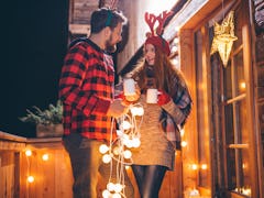 A couple dressed in festive clothing holds their holiday drinks while stringing up Christmas lights ...
