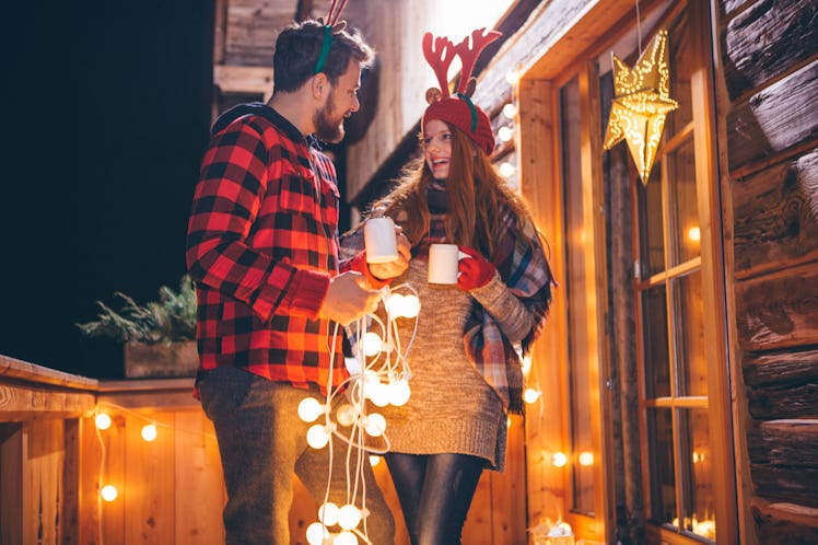 A couple dressed in festive clothing holds their holiday drinks while stringing up Christmas lights ...