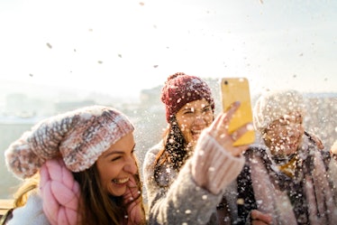 A group of women laugh while filming a snow day TikTok on a rooftop at golden hour.
