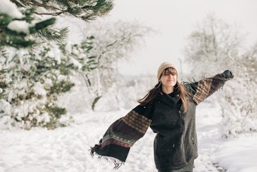 A young woman poses for a snow day TikTok in a snowy forest while wearing a winter outfit.