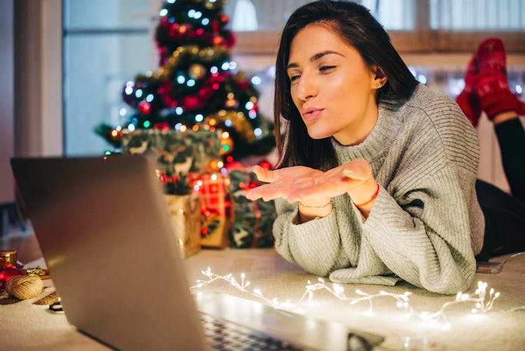 A girl lying down on fairy lights blows a kiss to her laptop screen during her virtual Christmas par...