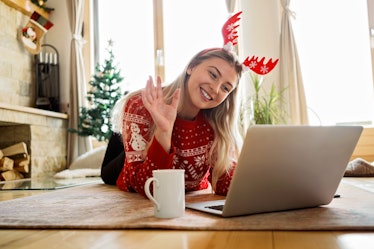 A woman in red reindeer antlers and a red sweater smiles at her virtual Christmas party on her lapto...