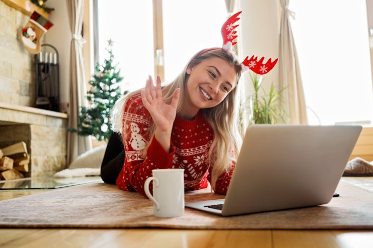A woman in red reindeer antlers and a red sweater smiles at her virtual Christmas party on her lapto...