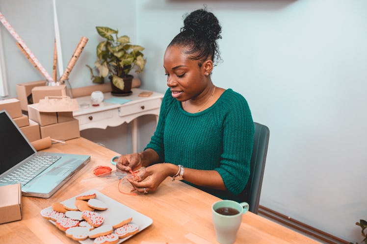 A young Black woman smiles while crafting gifts for the holidays and drinking tea.