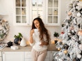 A woman enjoys her mug of coffee in a bright kitchen next to her decorated white Christmas tree.