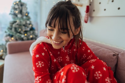 A happy woman wearing holiday pajamas sits on her couch next to her Christmas tree. 