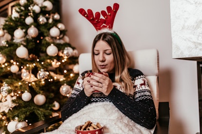 A woman in a holiday sweater sits in a chair next to her Christmas tree and blows on her drink.