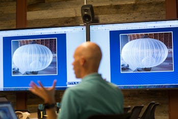 A man is seen walking past a screen depicting Loon balloons on two screens. The balloons are colored...