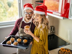 A man and woman in matching Santa hats bake cupcakes and other Christmas treats together in the kitc...