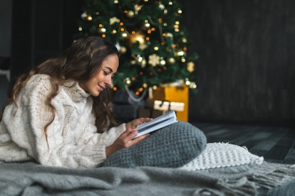 A happy woman reads a book on the floor next to a Christmas tree.