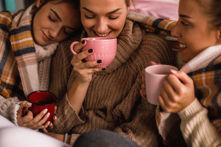 Three friends wearing sweaters, cuddle close while sipping on hot chocolate. 